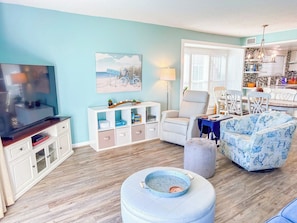 A living room with a television, a sofa, a dining area with chairs, and a kitchen in the background. - Beach Villas at Cherry Grove C (North Myrtle Beach)