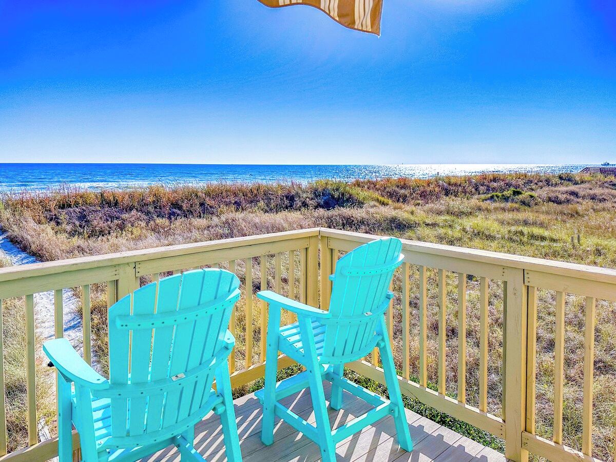 A wooden deck with two turquoise Adirondack chairs facing a beach.