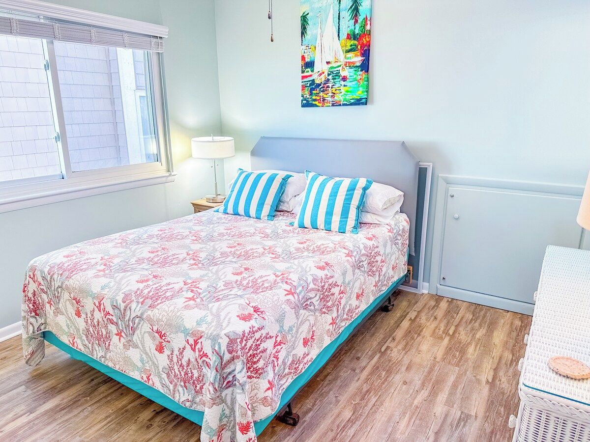 A bedroom with a bed, a nightstand, a lamp, a painting, and a wicker chair.