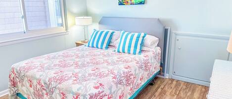 A bedroom with a bed, a nightstand, a lamp, a painting, and a wicker chair.