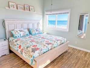 A bedroom with a bed, a nightstand, a dresser, a window with a view of the ocean, and a shelf with framed pictures. - Beach Villas at Cherry Grove C (North Myrtle Beach)
