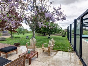 Une terrasse extérieure aménagée avec des meubles en bois, une table en pierre et une vue sur un jardin planté d’arbres en fleurs.