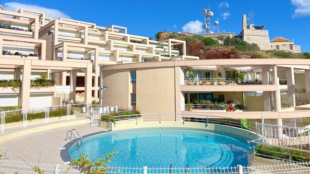 Ett modernt hotell med en swimmingpool, flera balkonger och utsikt över en historisk byggnad i bakgrunden.