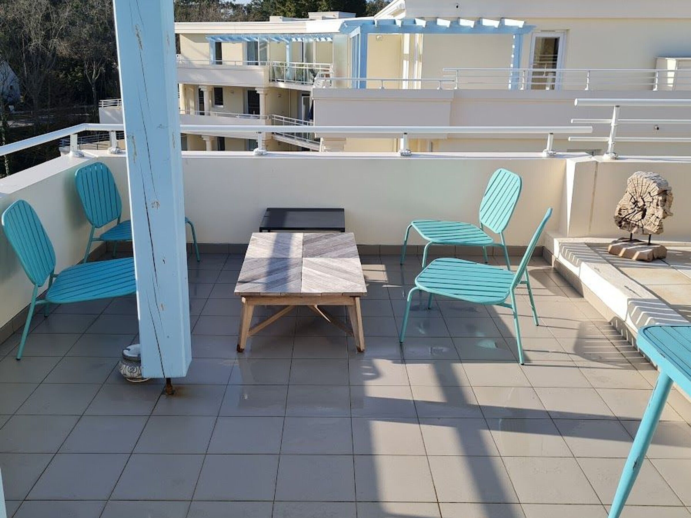 A rooftop terrace with a wooden table and two blue chairs, a small sculpture, and a view of a building in the background.
