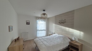 Ett sovrum med en stor säng, en byrå i trä, ett element, ett fönster med tunna gardiner, en hängande lampa och en väggmonterad lampa.