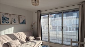 Ett modernt vardagsrum med en beige soffa, ett litet runt bord och ett stort fönster med tunna, genomskinliga gardiner.