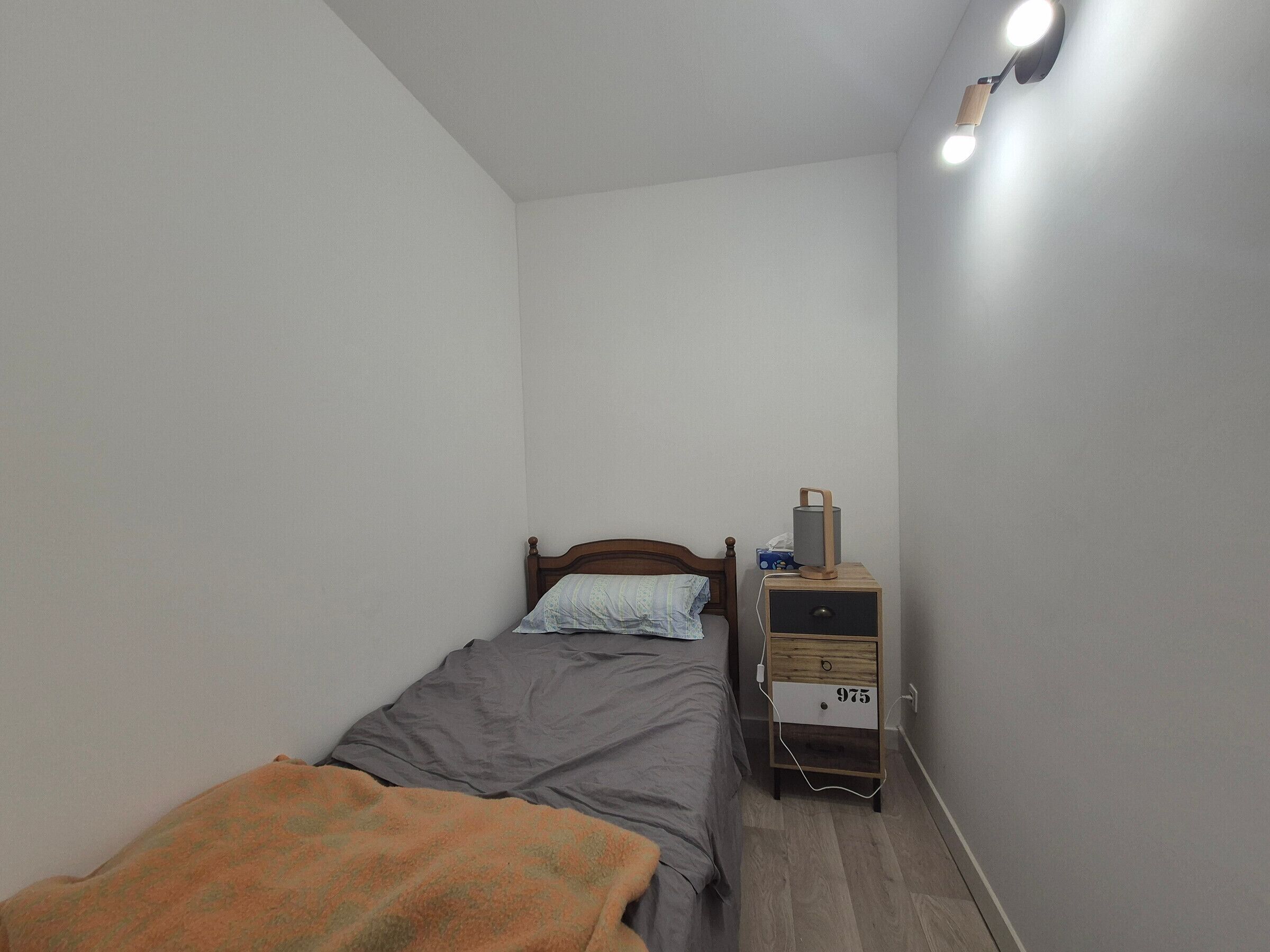 A small, neatly arranged bedroom with a single bed, a bedside table with a lamp, and a nightstand.