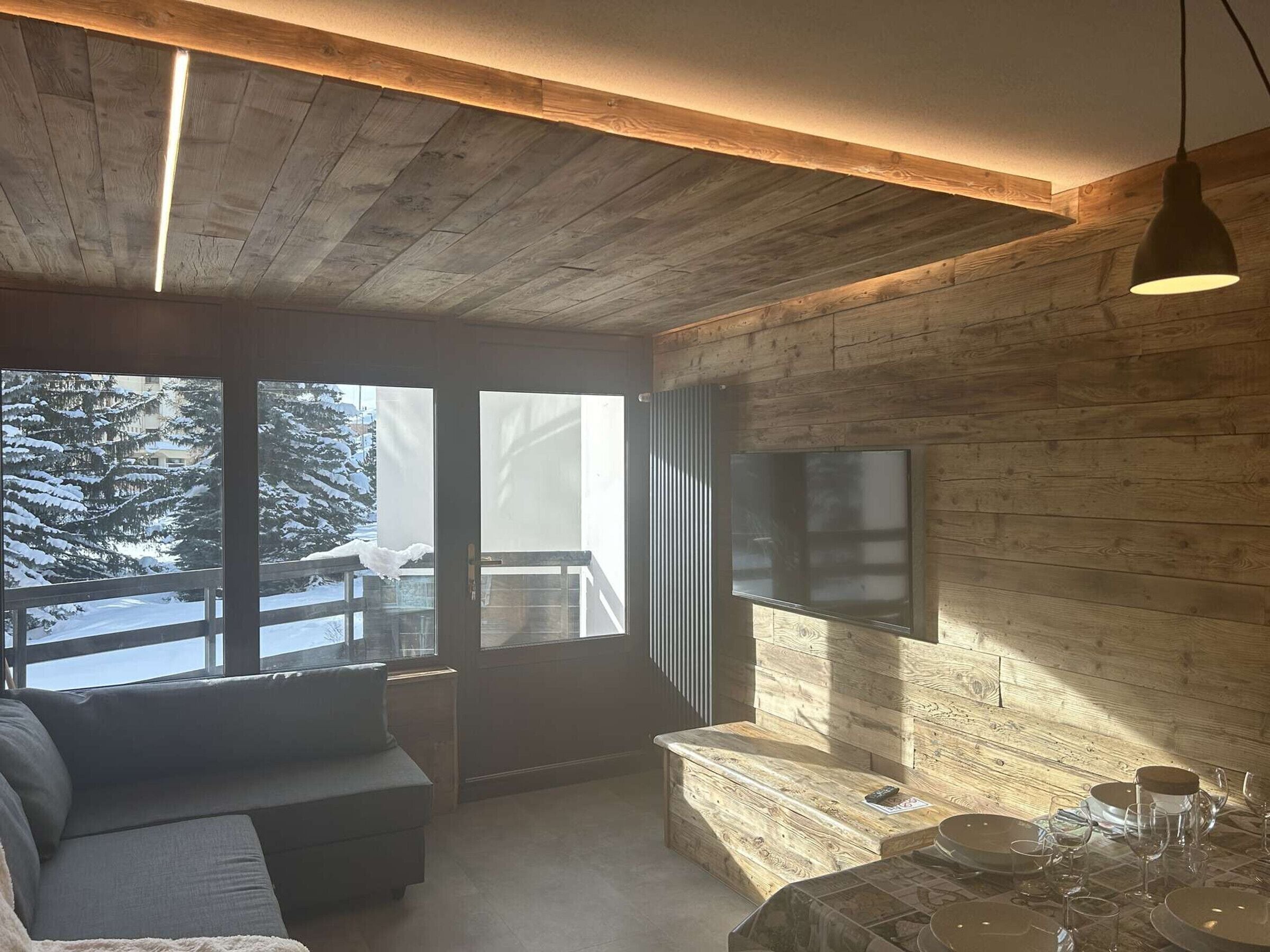 A cozy room with a wooden ceiling, a large window with a view of snow-covered trees, a flat-screen TV mounted on the wall, and a dining area with a table set for two.