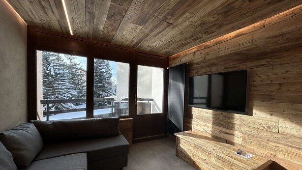 Un salon moderne doté d’un plafond en bois, de grandes fenêtres, d’un téléviseur encastré et d’un coin salon confortable.