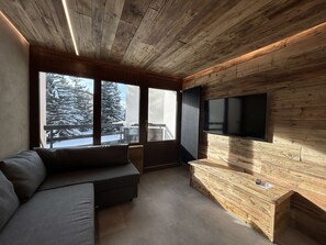 Ein modernes Wohnzimmer mit Holzboden, großen Fenstern, einem integrierten Fernseher und einer gemütlichen Sitzecke.