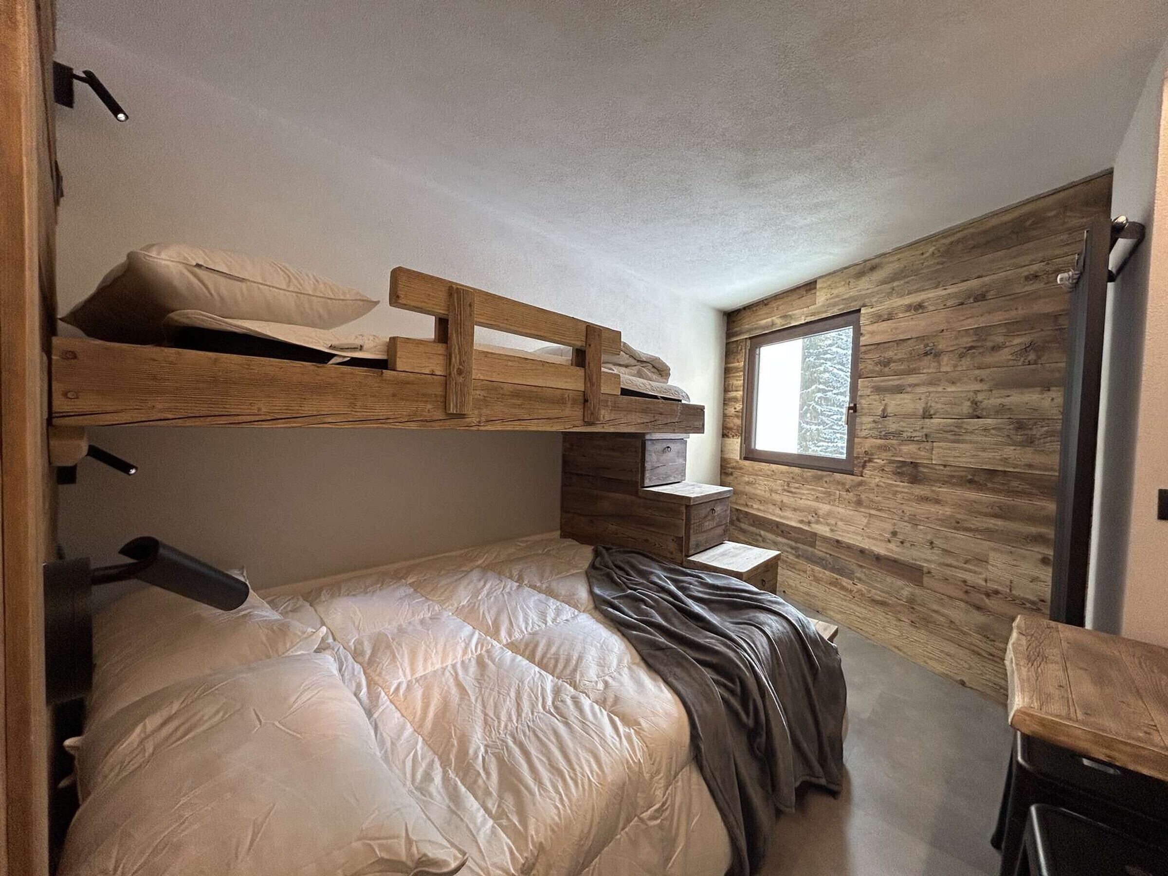 A compact bedroom with a bunk bed, a single bed, a small desk, and a window with a view of trees.