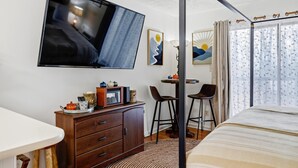 Hotellihuone, jossa on sänky, seinälle asennettu televisio, puinen lipasto, pieni pöytä tuoleineen ja ikkuna, jossa on verhot.
