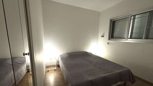 Una habitación pequeña y poco amueblada, con dos camas, una ventana con persianas y una puerta que da a otra habitación.
