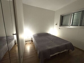 Ein kleiner, einfach eingerichteter Raum mit zwei Betten, einem Fenster mit Jalousien und einer Tür zu einem anderen Raum.