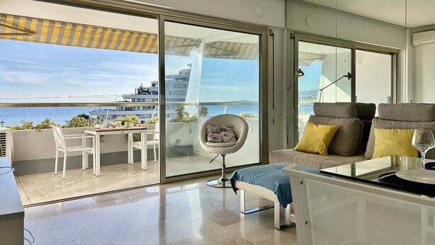 Un balcón moderno con vistas al mar y edificios, un área de descanso con cojines y una mesita con una lámpara.