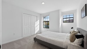Un dormitorio con cama, dos ventanas, una puerta y una mesita de noche con lámpara.