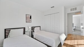 Quarto de hotel com duas camas de solteiro, uma mesa de cabeceira com abajur, uma escrivaninha de madeira e uma porta que dá para outro quarto.