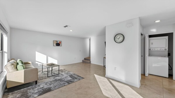 Uma sala de estar ampla com sofá, mesa de centro e tapete. Uma escada leva a outro andar e um relógio está visível na parede.