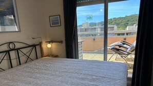 Een slaapkamer met een bed, een nachtkastje met een lamp, een foto in een lijst aan de muur, en uitzicht op het balkon met stoelen.