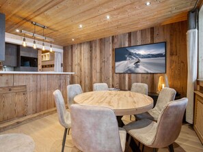 A modern kitchen with wooden paneling, a round dining table with six chairs, and a large TV on the wall. - Renovated ski-in ski-out apartment for 7 people (Courchevel 1850)