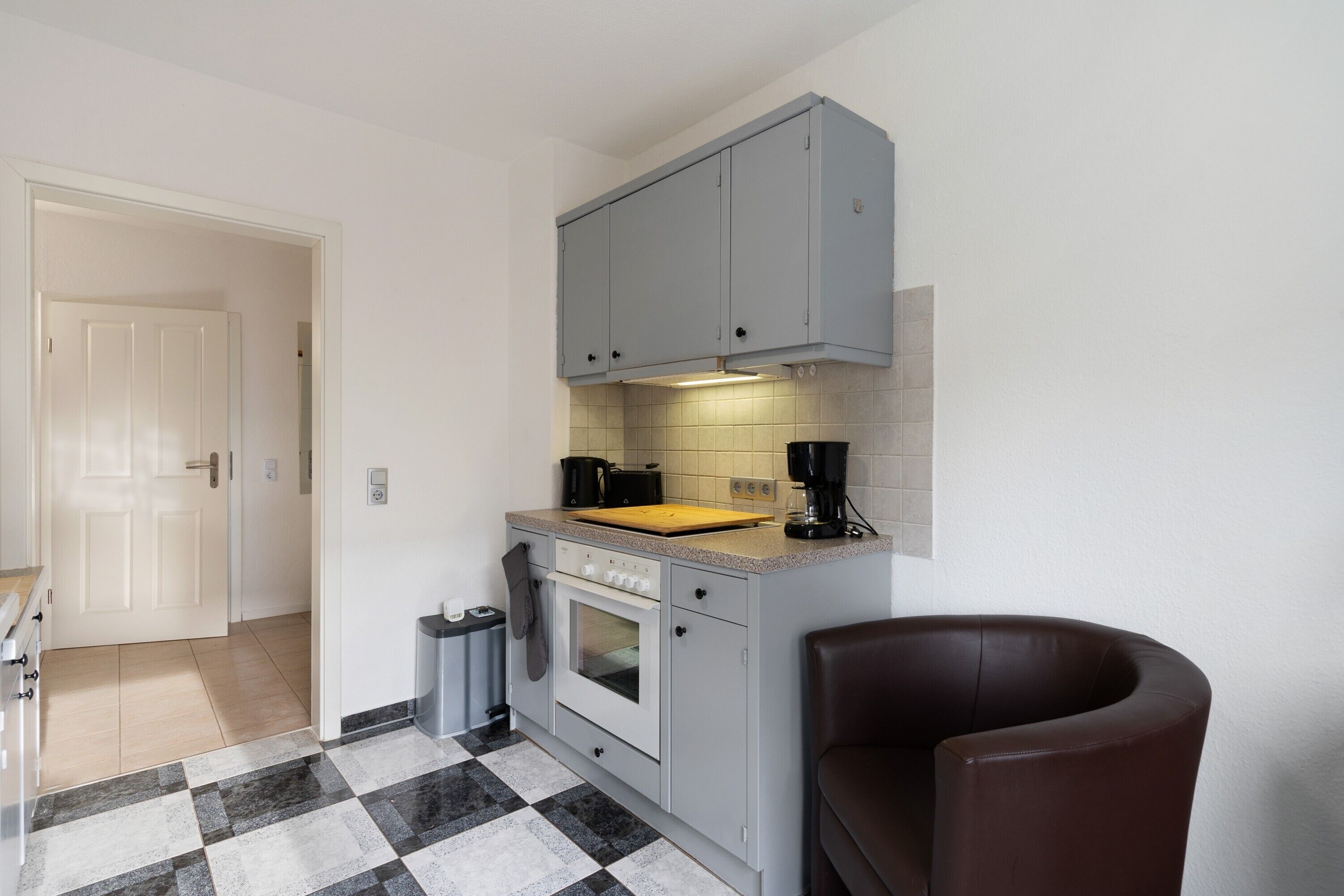 A compact kitchen with grey cabinets, a wooden countertop, a coffee machine, and a black trash bin.