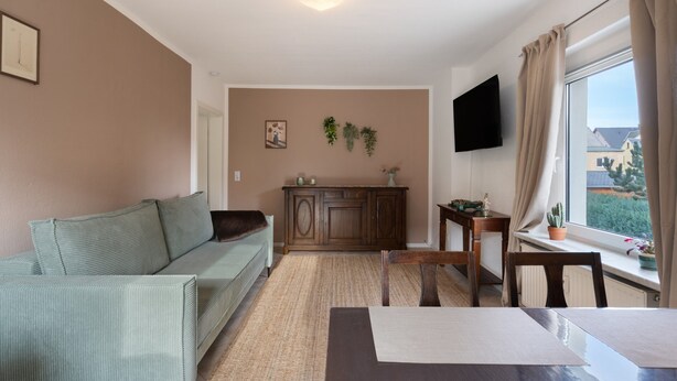 A living room with a sofa, a dining table, a TV mounted on the wall, and a window with curtains. - Apartment 'Yvette's 2' with Shared Garden and Wi-Fi (Lugau/Erzgeb.)