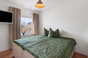 A bedroom with a bed, curtains, a TV, and a window with a view of buildings. - Apartment 'Yvette's 2' with Shared Garden and Wi-Fi (Lugau/Erzgeb.)