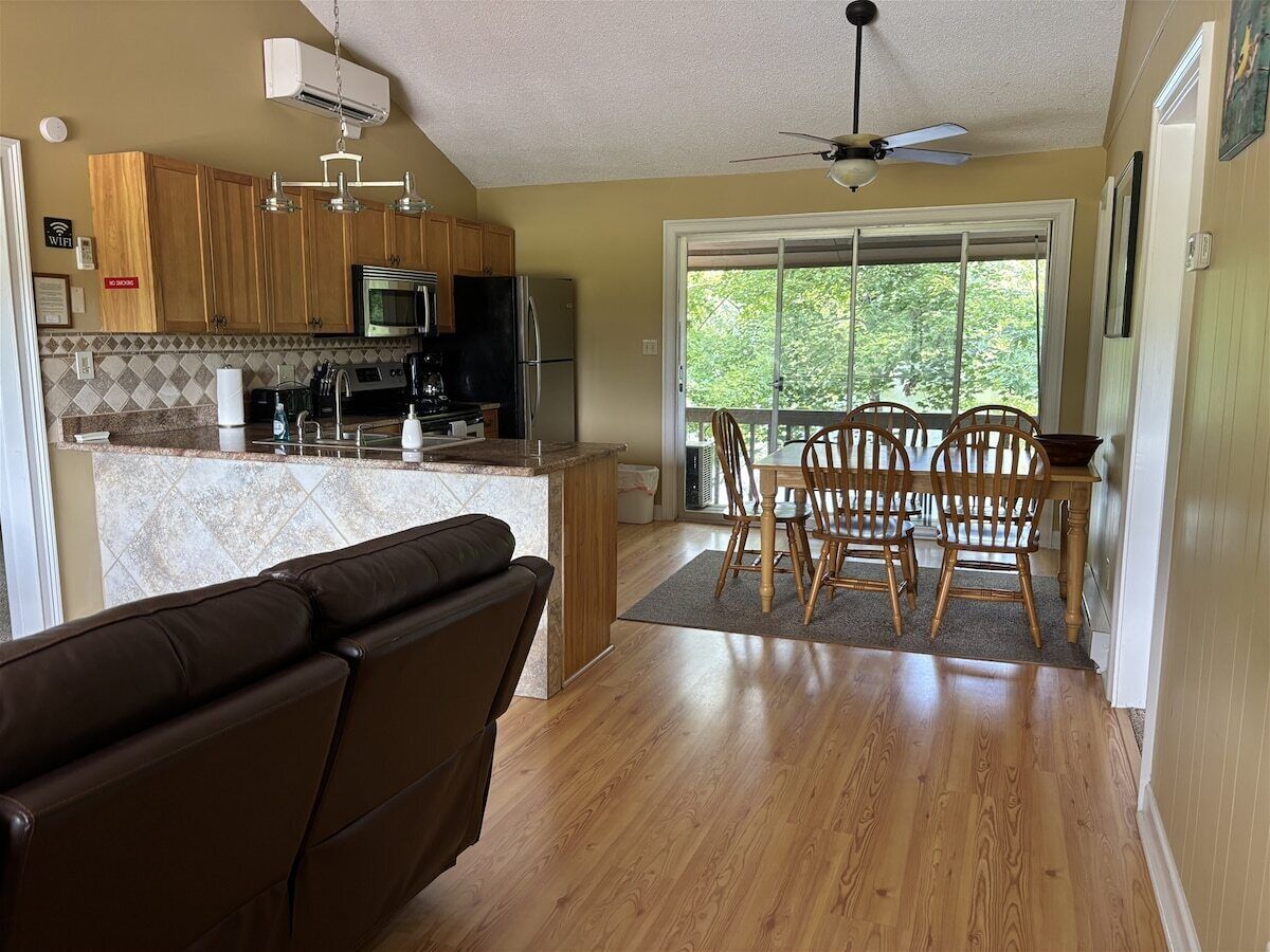 Une cuisine avec des meubles de rangement en bois, une table à manger avec des chaises, un ventilateur de plafond et une grande fenêtre.