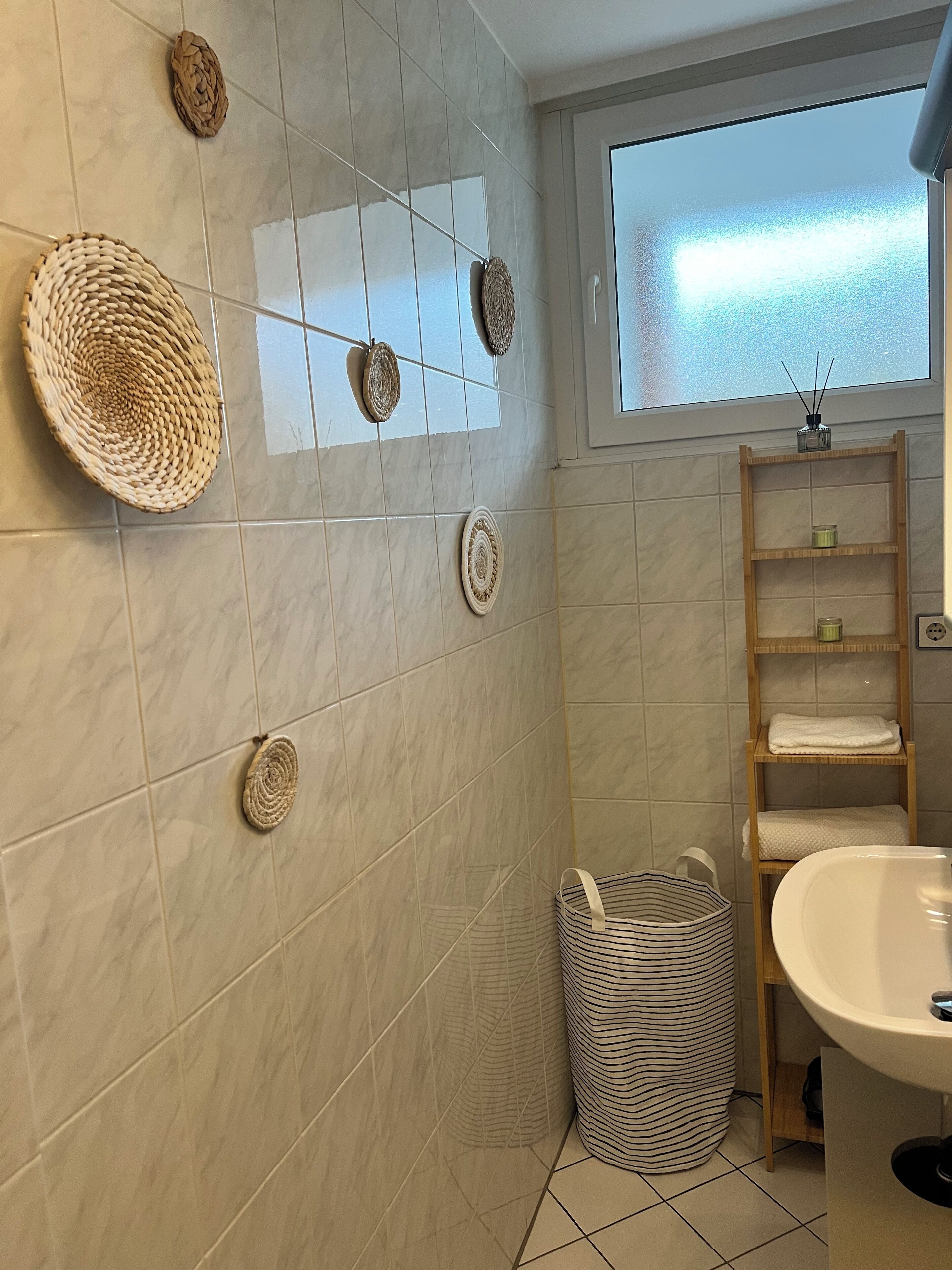 A bathroom with a tiled wall, a wooden shelf with towels, a striped trash bin, and woven wall decorations.