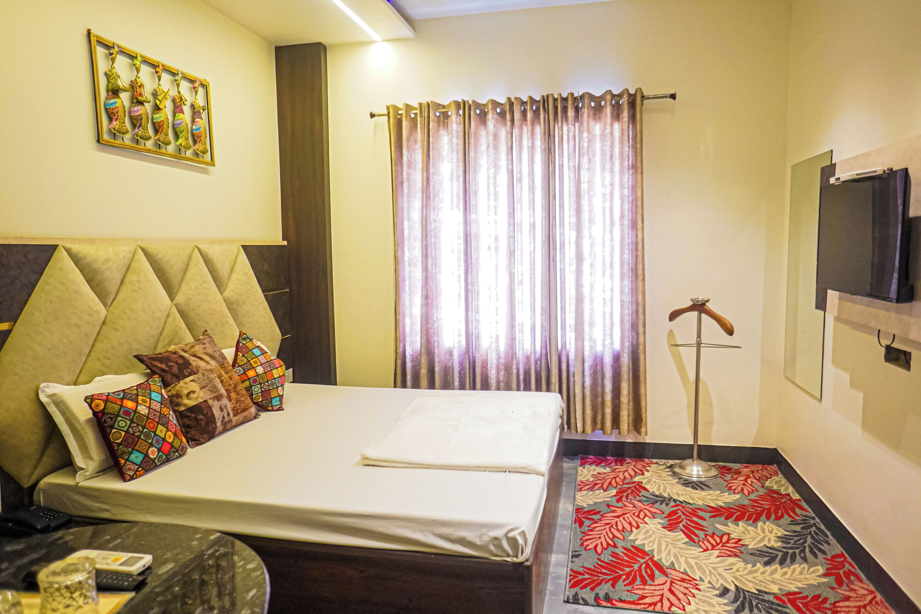 A hotel room with a bed, a TV mounted on the wall, a window with curtains, and a decorative wall piece.