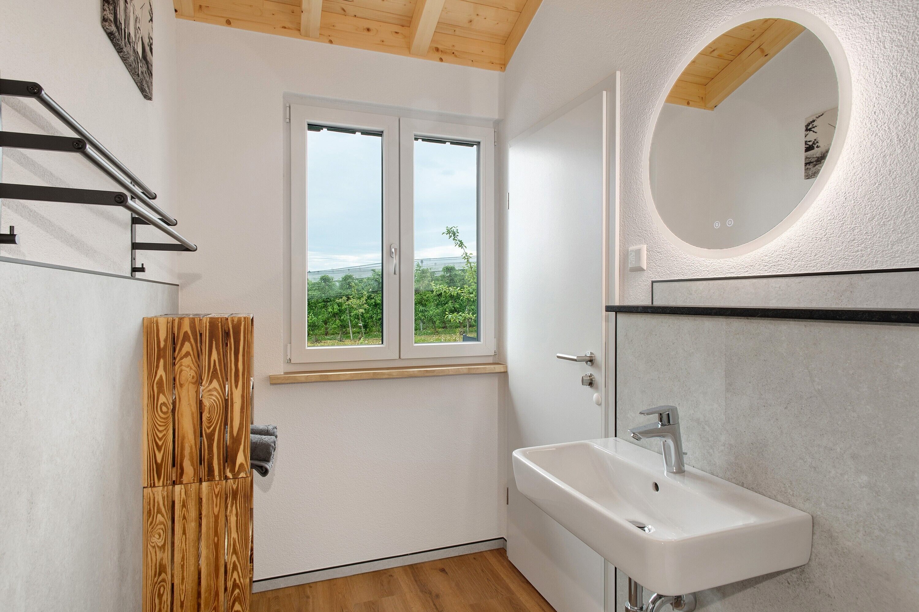 Een badkamer met een witte wastafel, een houten kastje, een raam met uitzicht en een ronde spiegel.