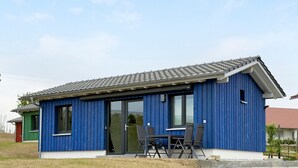 Un bâtiment bleu avec un toit recouvert de tuiles, de grandes portes coulissantes en verre et un petit patio agrémenté d’une table et de chaises.