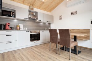 A modern kitchen with white cabinetry, a built-in oven, a microwave, and a coffee machine. There are two brown chairs and a wooden dining table. - Holiday Home 'Haus Kirsche' with Private Terrace, Wi-Fi & AC – Dog Friendly (Kressbronn am Bodensee)