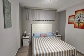 Une chambre à coucher comprenant un lit, deux tables de chevet, une commode, une fenêtre avec des rideaux et deux tableaux accrochés au mur.