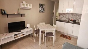 Un espacio habitable compacto con televisión, mesa de comedor, sillas, una zona de cocina y un estante pequeño.
