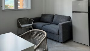 A compact living space with a grey sofa, a wicker chair, a white table, and a built-in refrigerator.