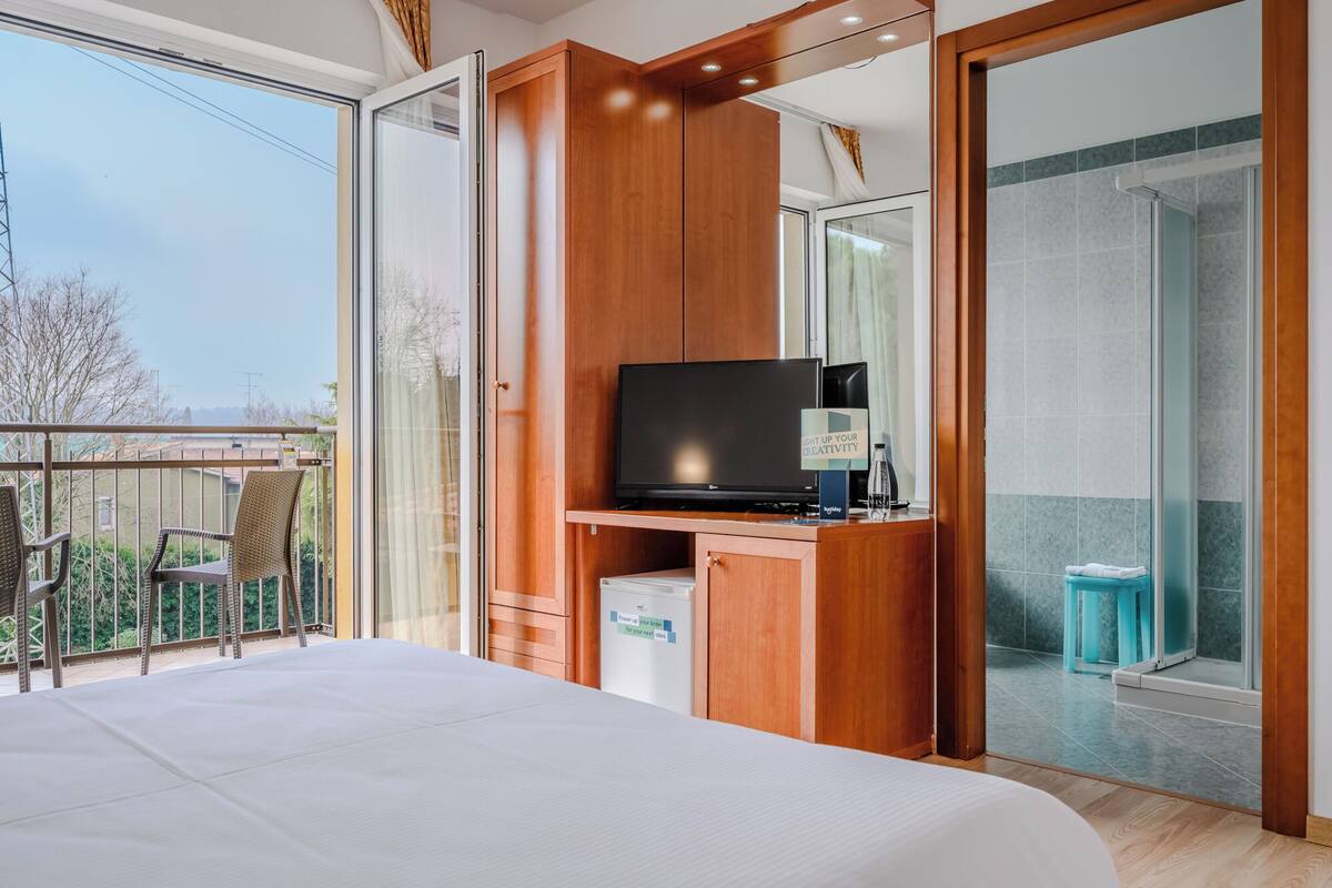 Habitación de hotel con cama, televisor, minibar y balcón con sillas.