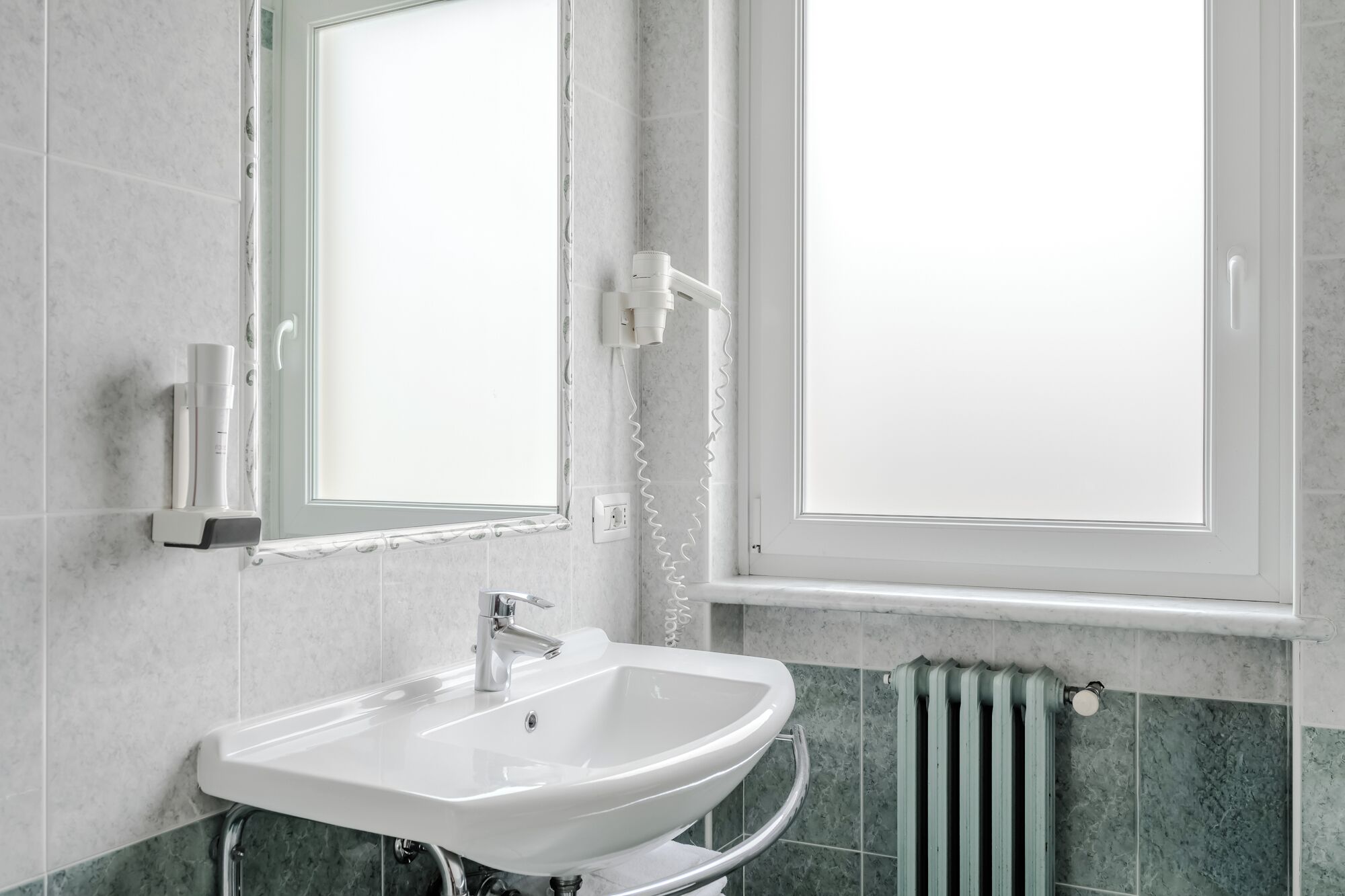 Une salle de bain avec un lavabo blanc, un miroir, une fenêtre et un radiateur.