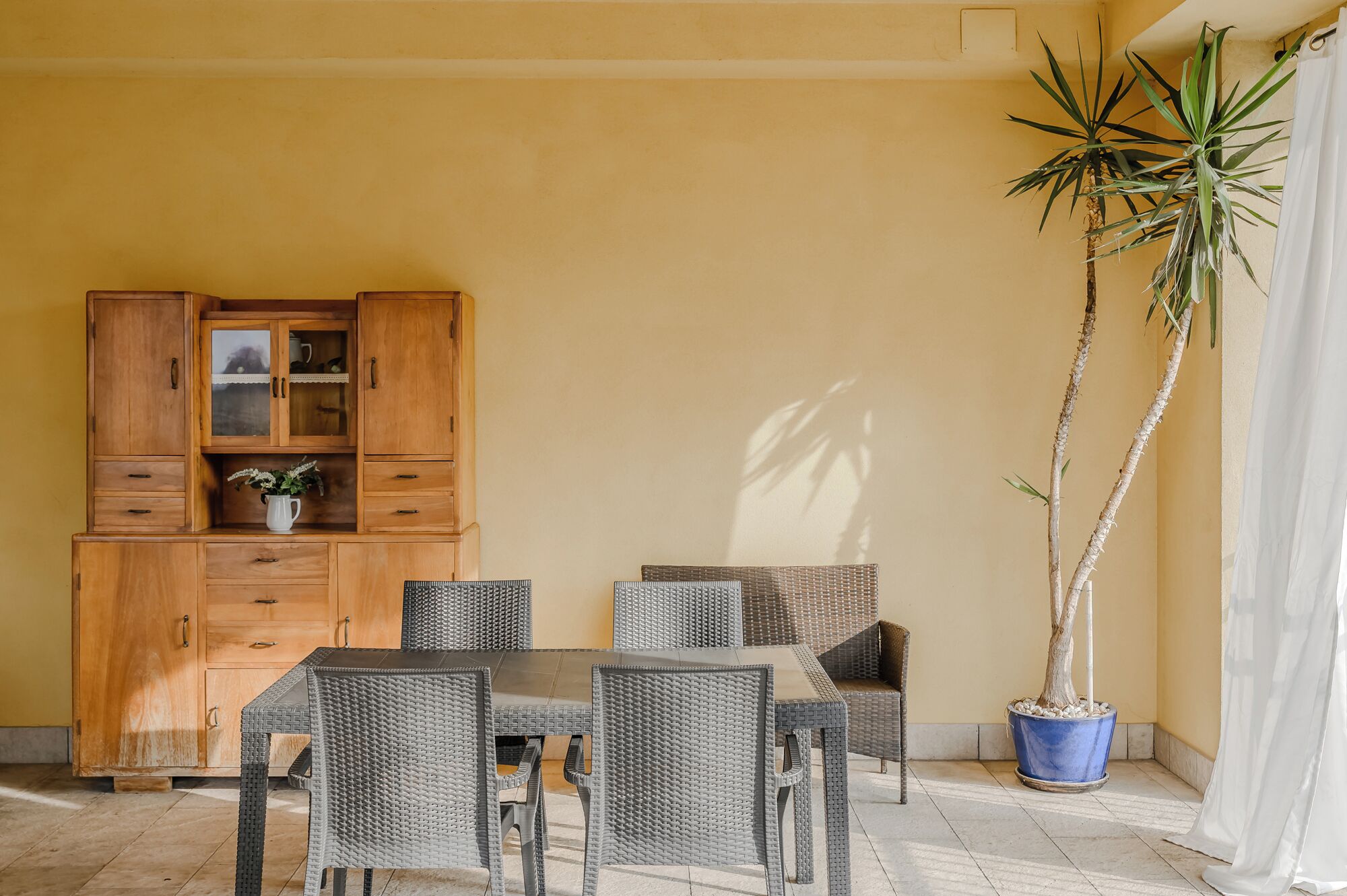 A dining area with a wooden cabinet, wicker chairs, a table, and a potted plant.