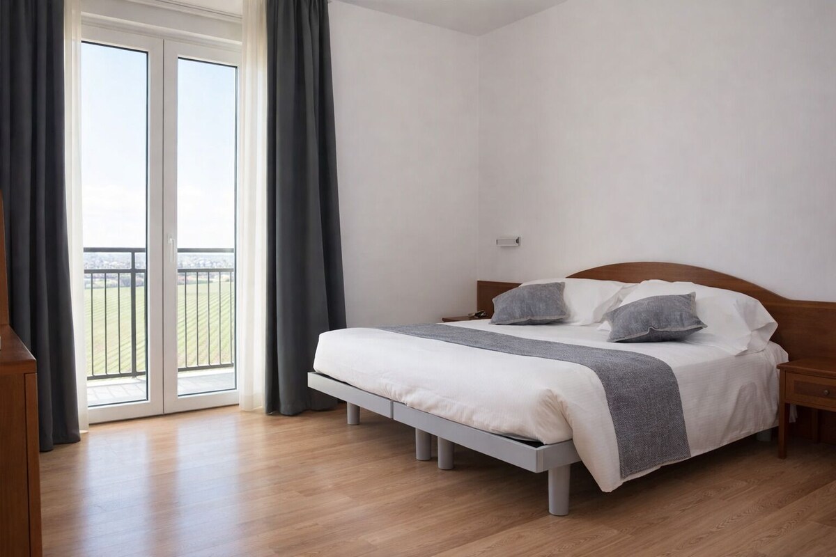 Ein Schlafzimmer mit Bett, Nachttisch, Fenster mit Vorhängen und Blick auf einen Balkon.