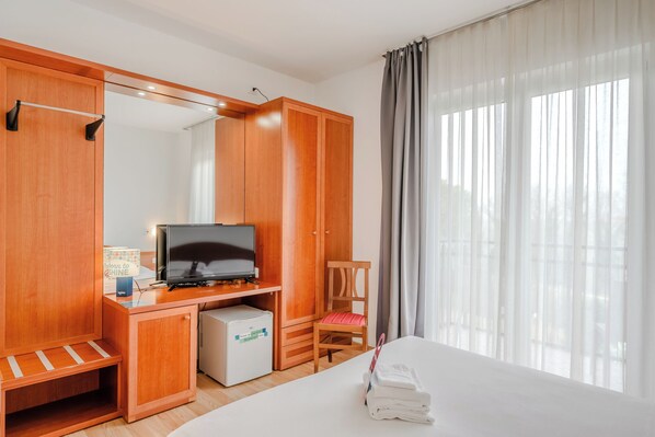A hotel room with a bed, a TV, a mini-fridge, a wooden wardrobe, a chair, and a large window with curtains. - Hotiday Peschiera Frassino (Broglie)