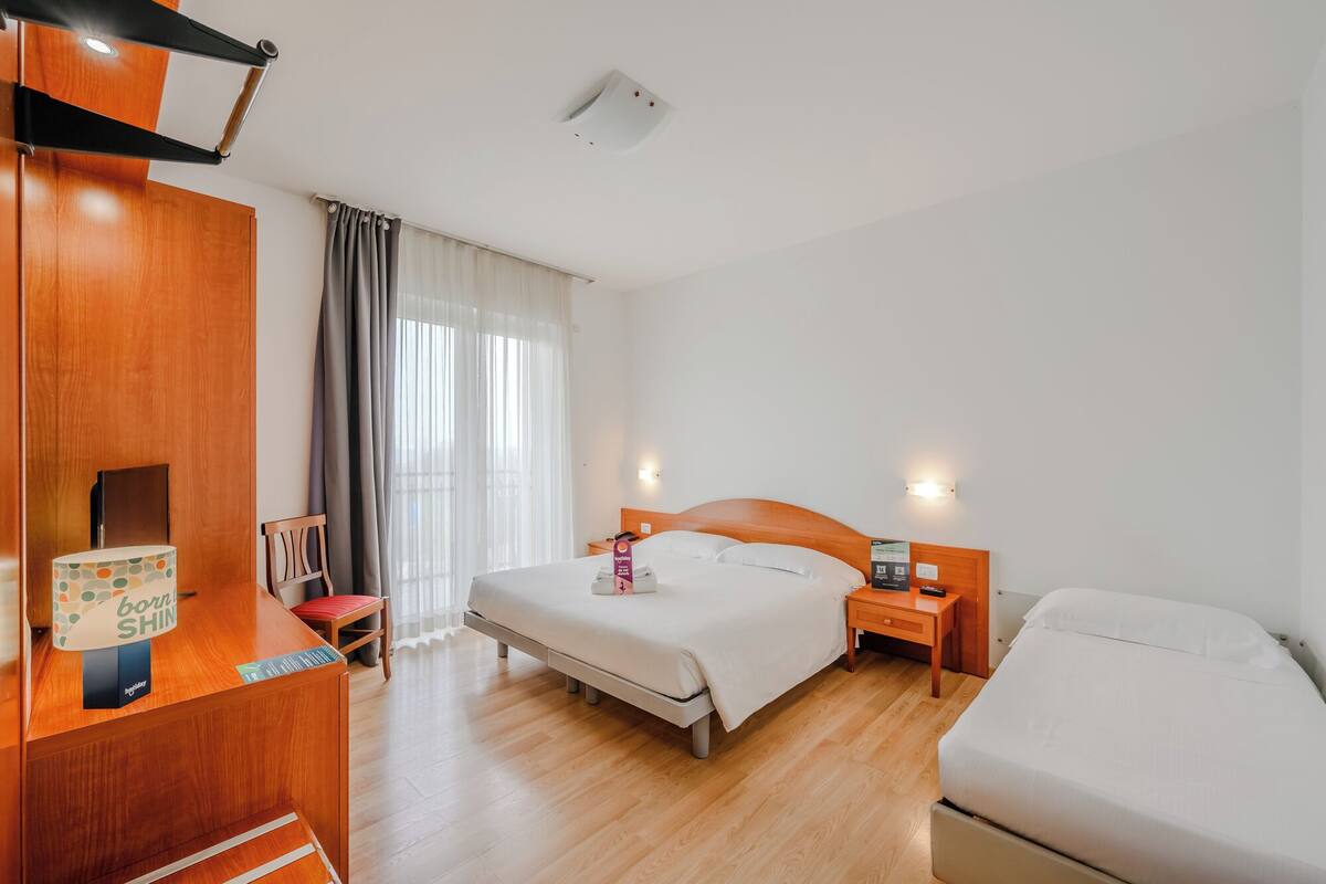 Una habitación de hotel con un armario de madera, un escritorio con lámpara, una cama con cabecera y una mesita de noche con lámpara.