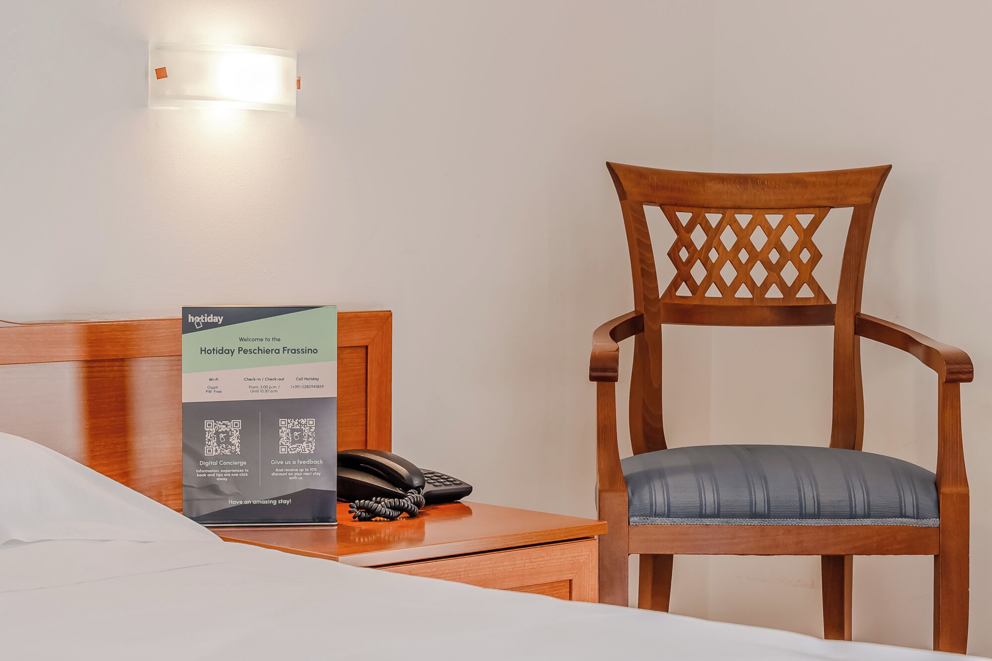 A hotel room with a wooden chair, a bedside table with a phone, and a signboard with hotel information.