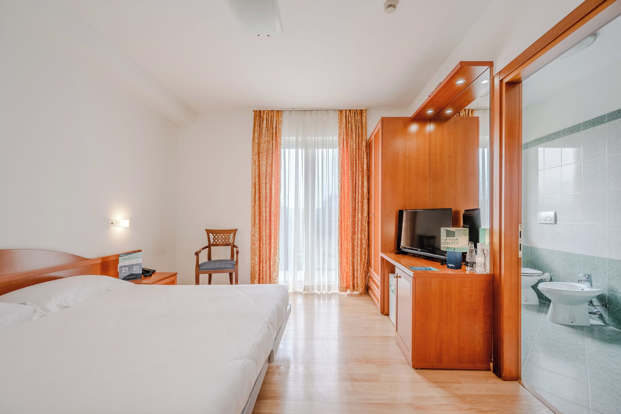 A hotel room with a bed, a desk with a computer, a chair, a television, and a bathroom with a sink and toilet.