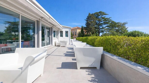 Un patio exterior moderno con mobiliario blanco, ventanas amplias y vistas al entorno verde.
