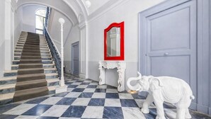 Un amplio pasillo con suelo de mármol a cuadros, una escalera con barandilla blanca, un espejo de marco rojo y una estatua de elefante blanco.