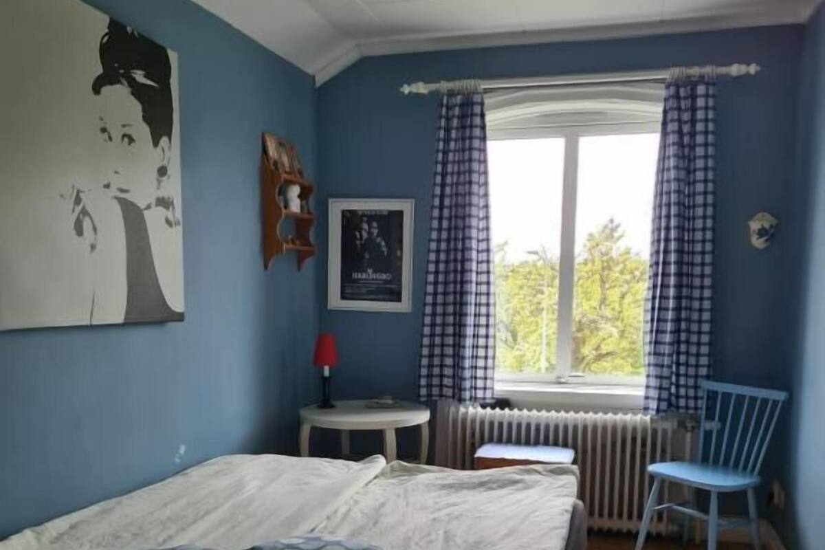 Un dormitorio con cama, ventana con cortinas, radiador y una silla.