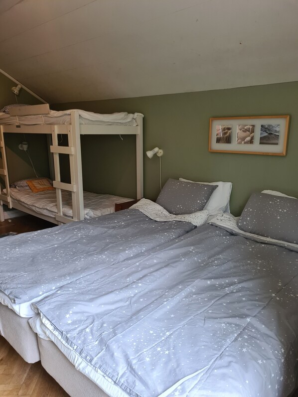 Uma cama de solteiro com escrivaninha e cadeira, mesa de cabeceira com abajur, e um quadro na parede.