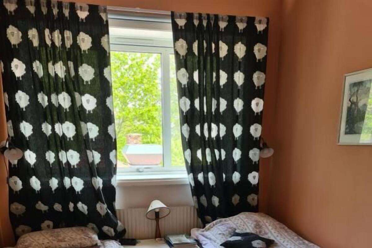 Un dormitorio con dos camas, una ventana con cortinas, una mesita de noche con lámpara y una alfombra a cuadros.