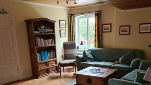 A cozy living room with a wooden floor, a green sofa, a bookshelf, and a window with curtains.